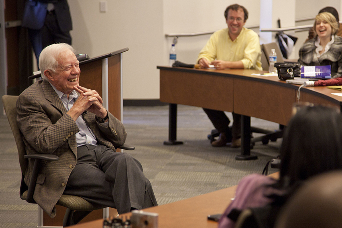 Former President Jimmy Carter sitting in chair smiling with college students in a classroom